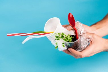 Mavi arka planda plastik nesneler ve yeşil fideleri tutan adamın kırpılmış görüntüsü