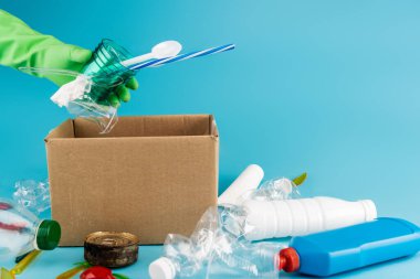 Lastik eldivenli temizleyicinin mavi arka plandaki karton kutuda plastik çöpleri toplaması.