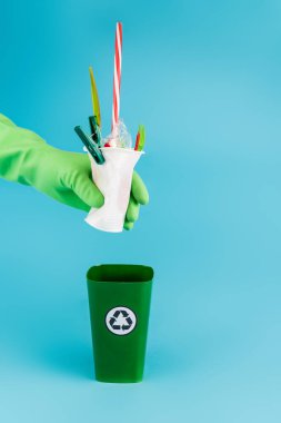 Lastik eldivenli temizleyicinin mavi arka plandaki çöp kutusuna plastik çöp atması, panoramik çekim