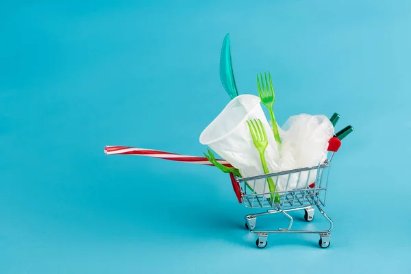 青の背景に小さなショッピングカートに使い捨てのプラスチックオブジェクト — ストック写真