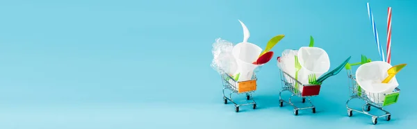 disposable plastic objects in small shopping carts on blue background, panoramic shot