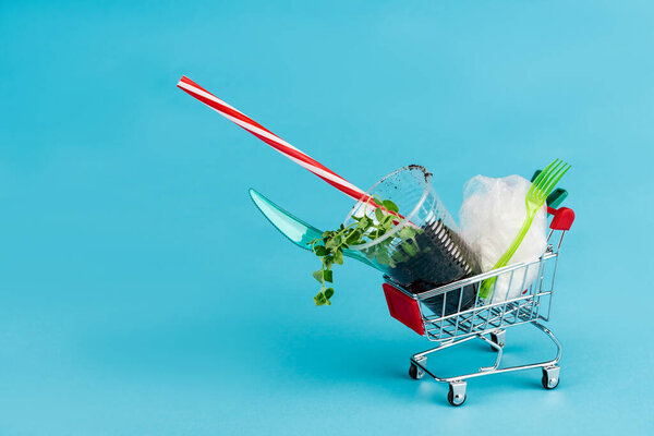 disposable plastic objects and plant in ground in small shopping cart on blue background