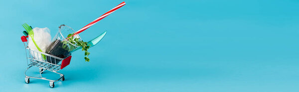 disposable plastic objects and plant in ground in small shopping cart on blue background, panoramic shot