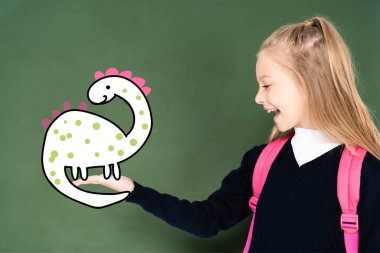 Okullu kız elinde yeşil karatahtadaki resimli dinozoru işaret ediyor. 