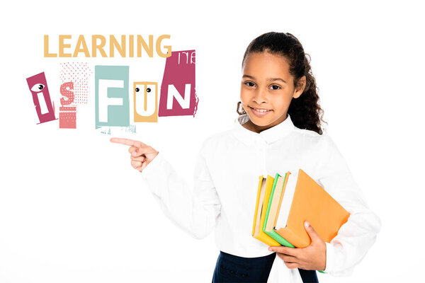 african american schoolgirl pointing with finger at learning is fun lettering while holding books isolated on white