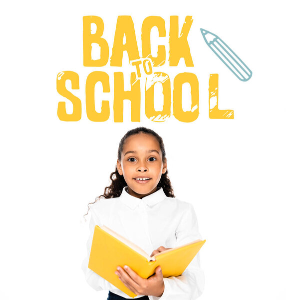 excited african american schoolgirl holding book and looking at camera isolated on white, back to school illustration