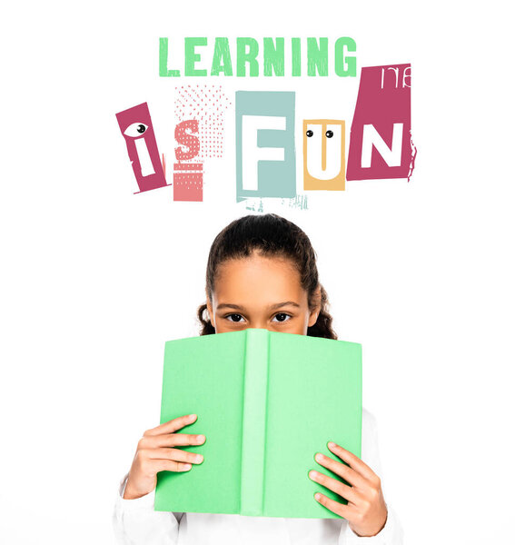 african american schoolgirl looking at camera while holding book near face isolated on white, learning is fun illustration