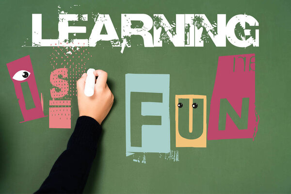 cropped view of schoolgirl writing with chalk learning is fun lettering on green chalkboard