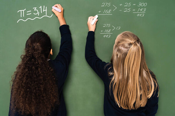 back view of multicultural schoolgirls writing mathematical equations on chalkboard