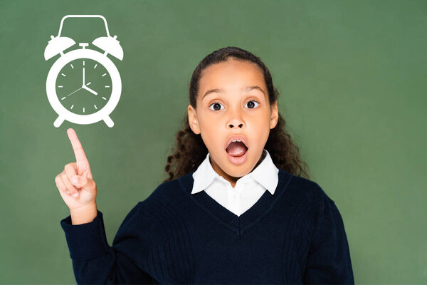 шокированная африканская школьница, указывающая на иллюстрированный будильник на зеленой доске