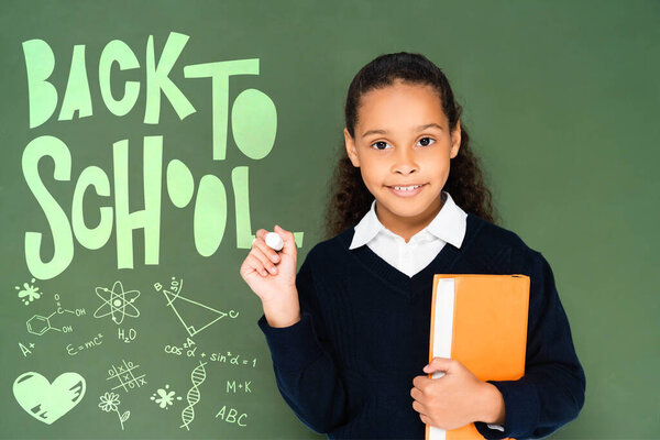 Африканская американская школьница держит кусок мела и книги, стоя рядом с доской со школьными надписями