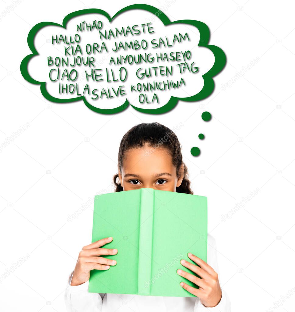 african american schoolgirl looking at camera while holding book near face isolated on white, illustrated speech bubble with languages
