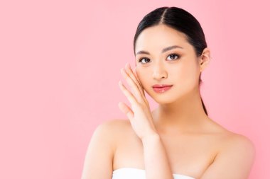 young asian woman looking at camera and touching face isolated on pink clipart