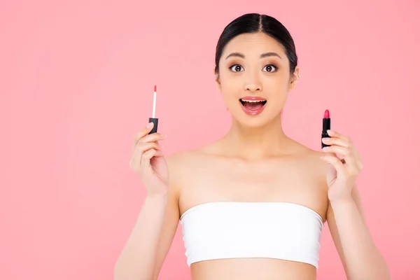 兴奋的亚洲女人一边拿着唇彩和唇膏 一边看着相机 用粉色隔离起来 — 图库照片
