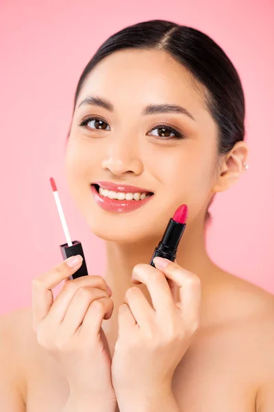 年轻的亚洲女人一边拿着唇彩和唇膏 一边看着相机 唇膏与粉红隔离在一起 — 图库照片