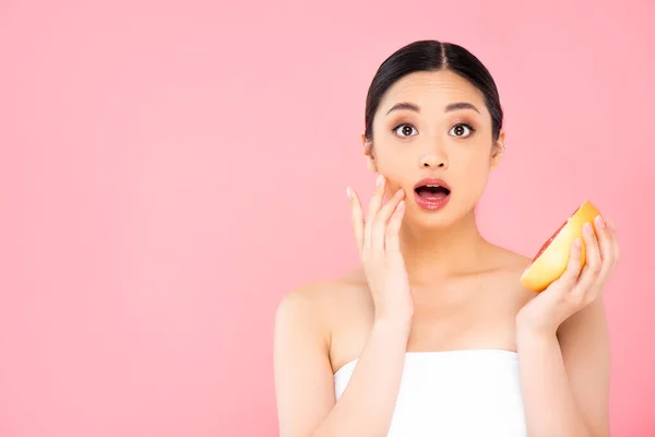 shocked asian woman touching face while holding half of grapefruit isolated on pink