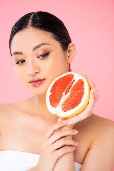 asian woman holding half of ripe grapefruit near face isolated on pink