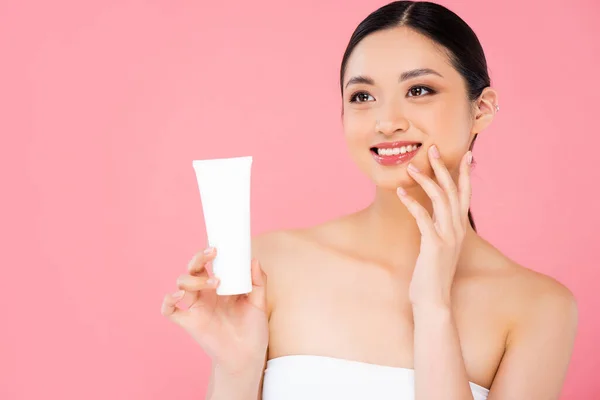 brunette asian woman holding tube of cosmetic cream, touching face and looking away isolated on pink