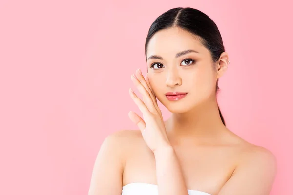 stock image young asian woman looking at camera and touching face isolated on pink