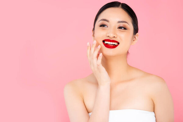 young asian woman with red lipstick on lips touching face while looking away isolated on pink