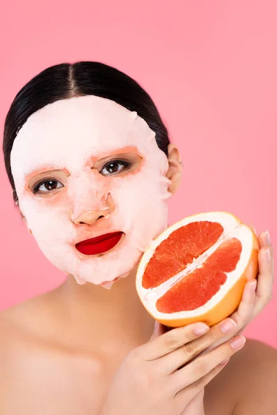 ピンク色に隔離されたカメラを見ながら新鮮なグレープフルーツの半分を持つ顔のマスクの若いアジア人女性 — ストック写真