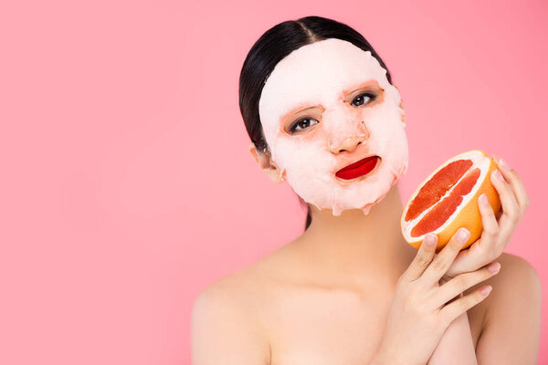 brunette asian woman in face mask holding half of ripe grapefruit while looking at camera isolated on pink