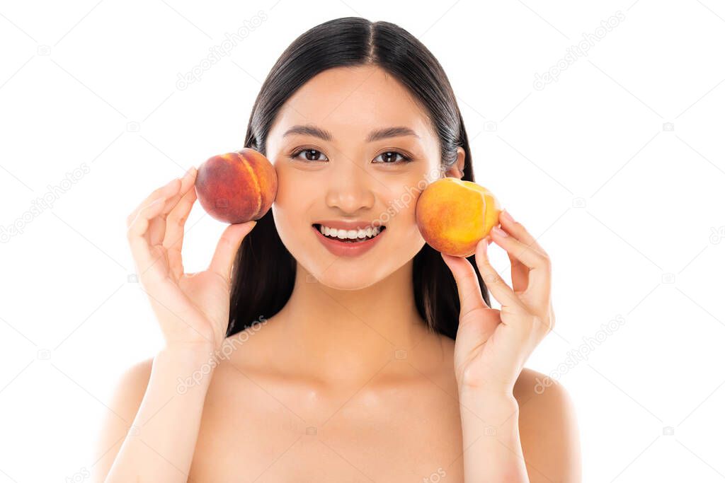 nude asian woman holding ripe peaches near face while looking at camera isolated on white