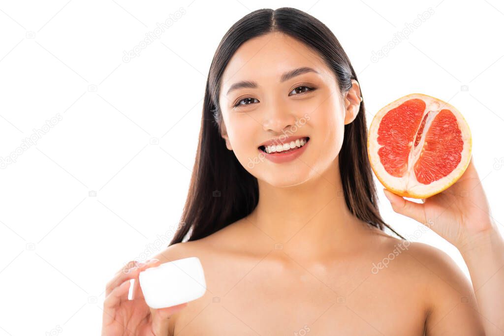 nude asian woman holding half of grapefruit and cosmetic cream while looking at camera isolated on white