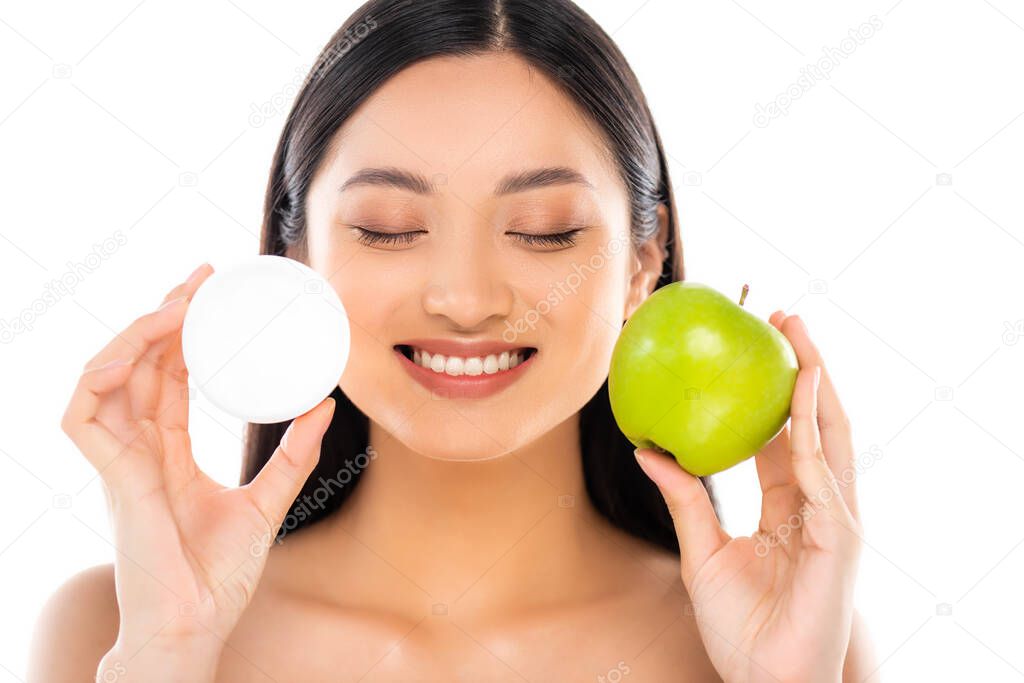 naked asian woman with closed eyes holding whole green apple and cosmetic cream near face isolated on white