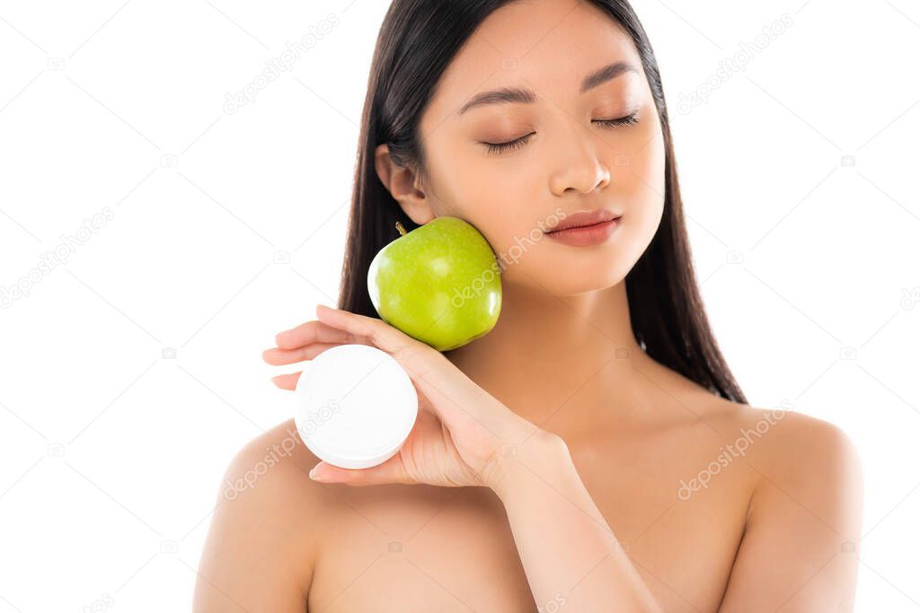 naked asian woman with closed eyes holding ripe apple and cosmetic cream near face isolated on white