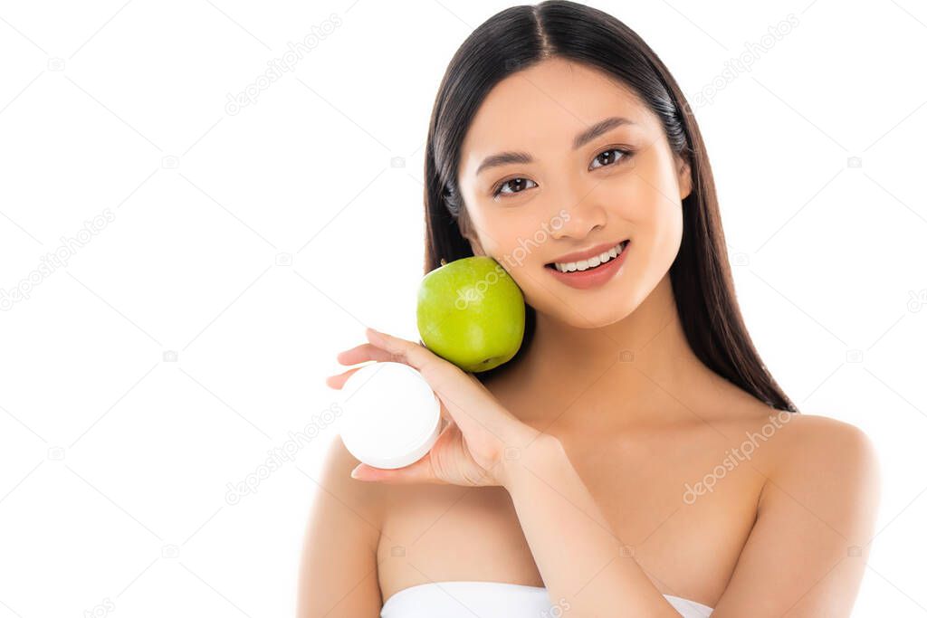 brunette asian woman holding green apple and cosmetic cream near face isolated on white
