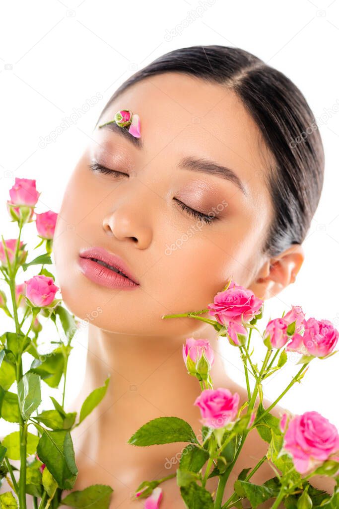 sensual asian woman with closed eyes and floral decoration on face near tiny roses isolated on white