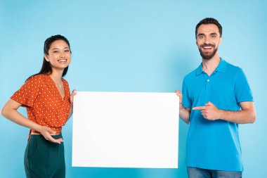 Polo tişörtlü sakallı bir adam ve kırmızı noktalı bluzlu Asyalı bir kadın mavi üzerinde boş bir levha taşıyor.