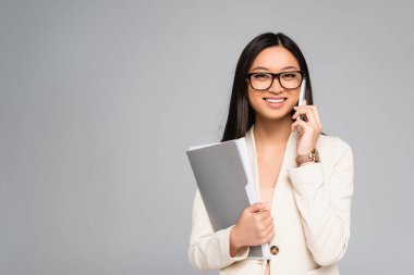 Beyaz ceketli, gözlüklü şık Asyalı iş kadını akıllı telefondan konuşuyor ve klasörü gri renkte tutuyor.