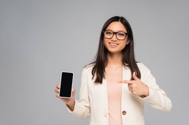 Gözlüklü genç Asyalı iş kadını gri ekranda boş ekranlı akıllı telefonu işaret ediyor.