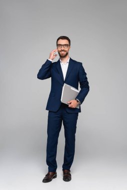 Mavi takım elbiseli genç işadamı, gri renkli cep telefonuyla konuşurken elinde belgeler tutuyor.