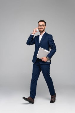 Mavi takım elbiseli iş adamı akıllı telefondan konuşuyor ve gri üzerinde yürürken klasörü tutuyor.