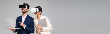 İş adamının gri üzerine izole edilmiş VR kulaklıkları kullanırken bilgisayarı işaret ettiği yatay görüntü