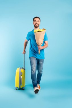 Polo tişörtlü ve kot pantolonlu genç turist elinde sarı buketle mavi bavulla yürürken...