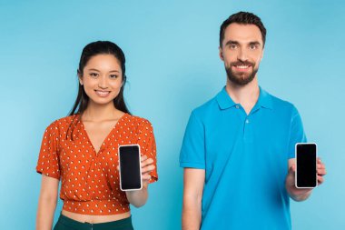 Kırmızı bluzlu Asyalı kadın ve polo tişörtlü adam mavi ekranlı akıllı telefonları gösteriyor.