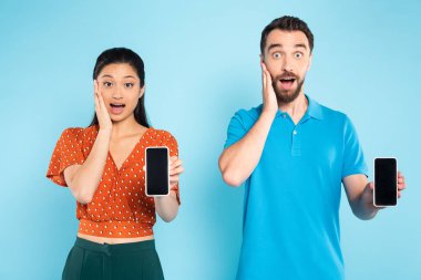 Mavi ekranda boş ekranlı akıllı telefonları tutarken yüzlerine dokunan ırklar arası şok edici bir çift.
