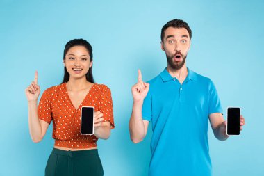 Genç Asyalı kadın ve heyecanlı adam mavi ekranda boş ekranlı akıllı telefonları tutarken fikir işareti gösteriyor.