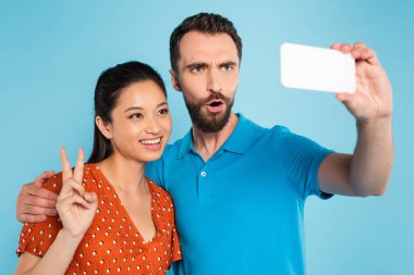 Asyalı bir kadınla selfie çekerken yüzünü buruşturan sakallı bir adam zafer jesti yapıyor.