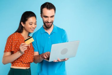brunette asian woman in red blouse holding credit card near man in polo t-shirt using laptop on blue clipart
