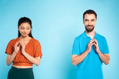 skeptical asian woman in red blouse and sly man in polo t-shirt gesturing with joined fingers on blue clipart