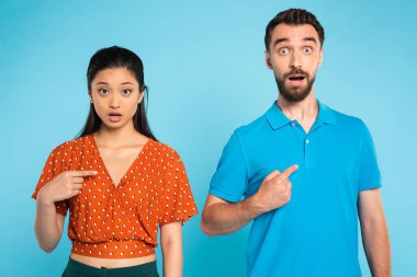 shocked asian woman in red blouse and bearded man in polo t-shirt pointing with fingers at themselves on blue clipart