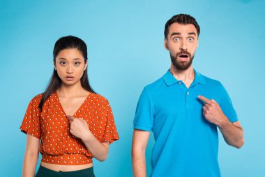surprised asian woman in red blouse and bearded man in polo t-shirt pointing with fingers at themselves on blue clipart