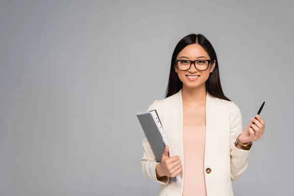 stylish asian businesswoman in white blazer and eyeglasses holding pen and folder isolated on grey