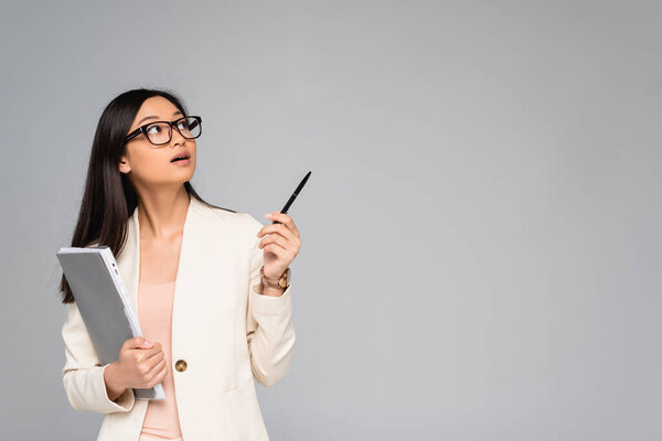 surprised asian businesswoman in white blazer looking away and pointing with pen isolated on grey