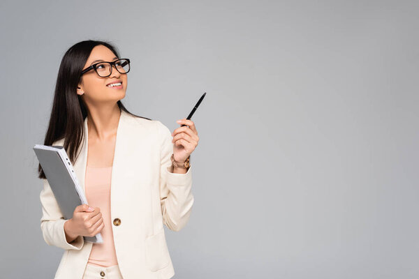 stylish asian businesswoman in white blazer and eyeglasses looking away and pointing with pen isolated on grey